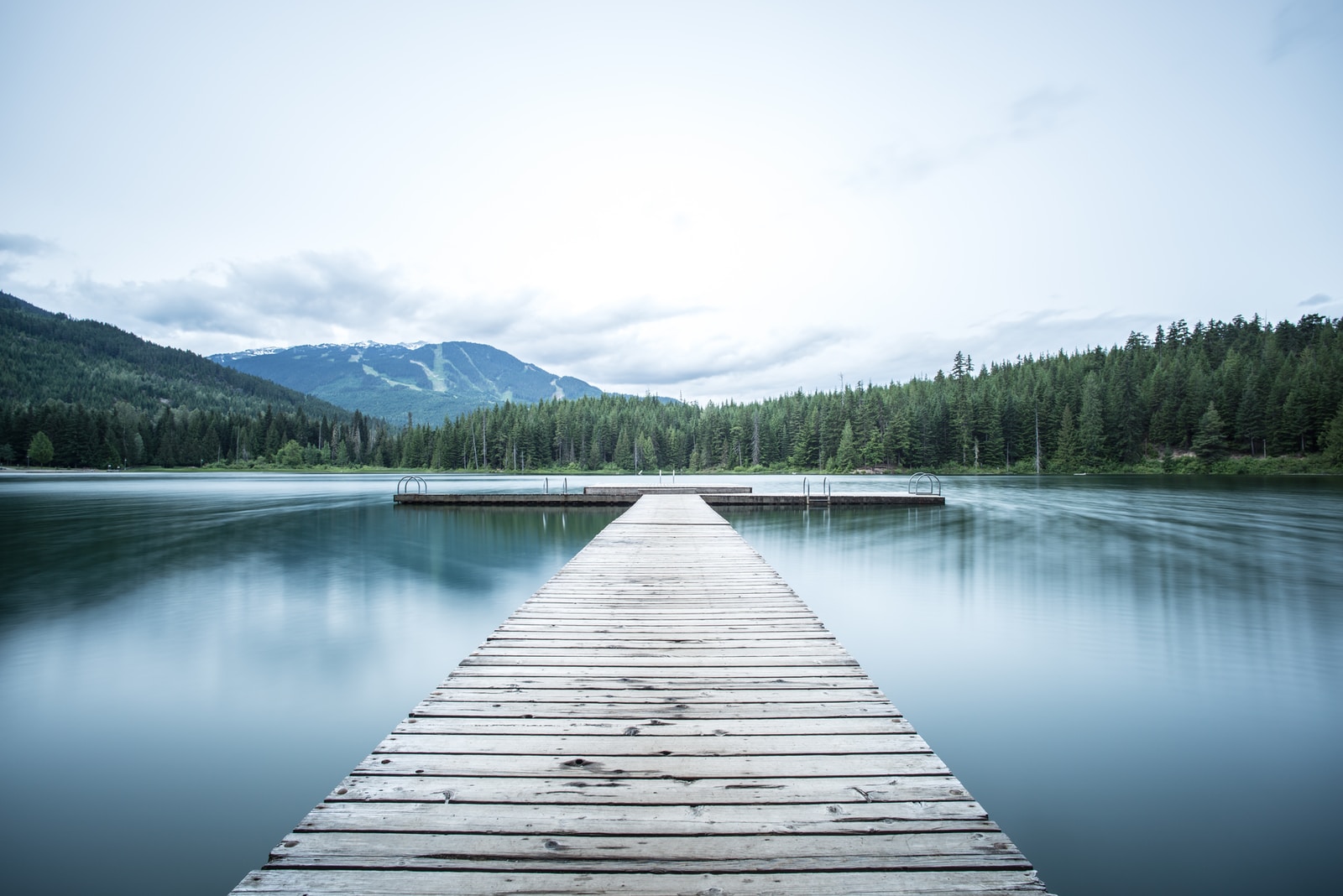 A long wooden dock made of slats of wood going outward to a horizontal dock with water on each side and trees and a mountain in the distance.